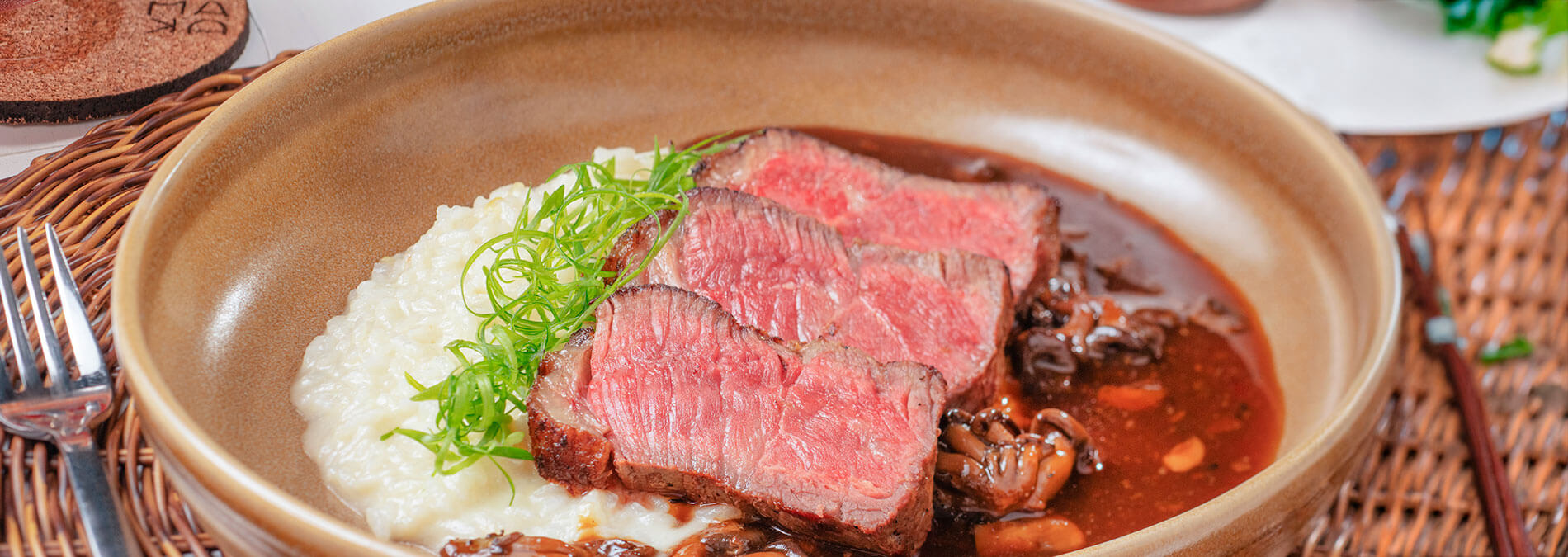 Prato com cortes de carne, purê e molho, servido em uma tigela de cerâmica e talheres no canto esquerdo. Uma mão segura um copo com um coquetel ao lado.