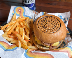  Um hambúrguer gourmet com o logo do impresso no pão, servido com batata frita em uma cesta. Uma lata de Pepsi está ao lado, o bucket oficial do resort ao fundo junto com outra pessoa comendo a mesma coisa. 
