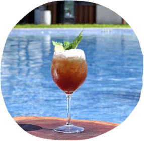 Um coquetel sofisticado em uma taça, com espuma e folhas de hortelã, em uma mesa de madeira ao lado da piscina do Japaratinga Lounge Resort.