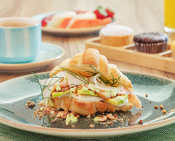 Um croissant recheado com proteína, frios, molhos e maçã verde em um prato de cerâmica. Em segundo plano, uma xícara de café, doces, frutas e um suco no buffet de café da manhã.