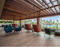 Vista da área externa do Moenda Café, com mesas, cadeiras e balanços suspensos sob uma pérgola de madeira com vista pra uma piscina e grandes coqueirais. 
