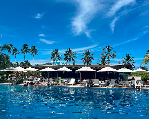 Vista da piscina principal do Japaratinga Lounge Resort com o Bar Verano ao fundo. Os hóspedes estão na piscina e nas espreguiçadeiras num dia com poucas nuvens.