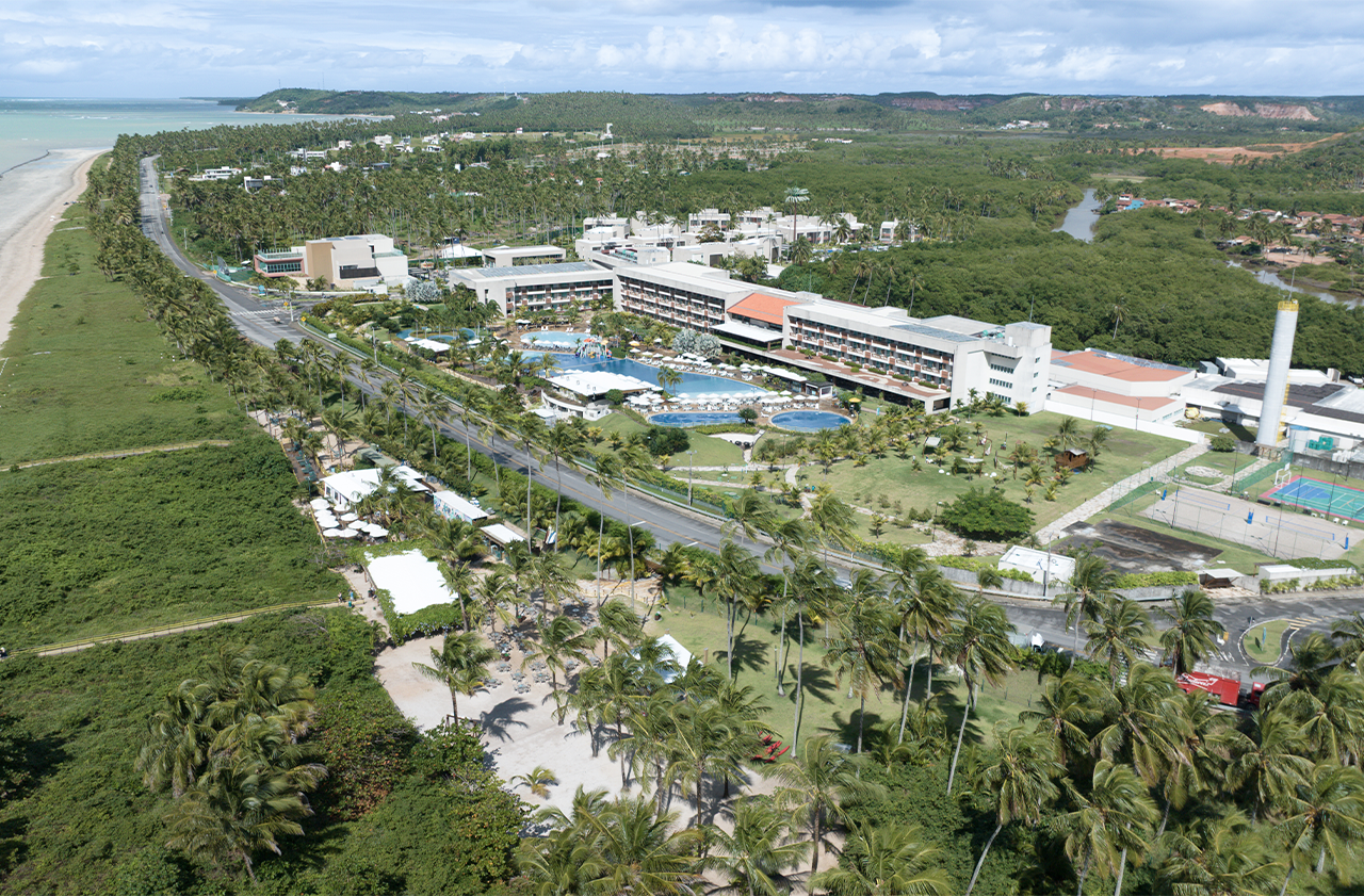 Vista aérea do Japaratinga Lounge Resort, mostrando todo a estrutura, piscinas principais e grande área verde de descanso. O resort é cercado por vegetação. É possível ver a pista que divide o resort da praia, e do Vila da Praia, o bar perto da areia da praia, que está com maré baixa, e areia clara.