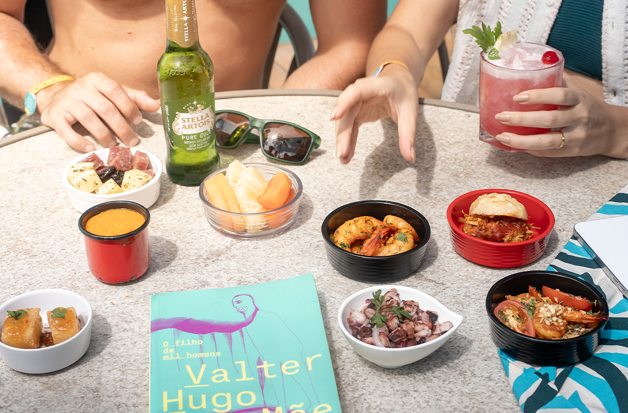 Uma mesa com petiscos (camarão, polvo, queijos e frutas) e drinks sendo a cerveja Stella Artois e um drink, sendo degustados por um casal à beira da piscina com um livro de cada azul no canto.