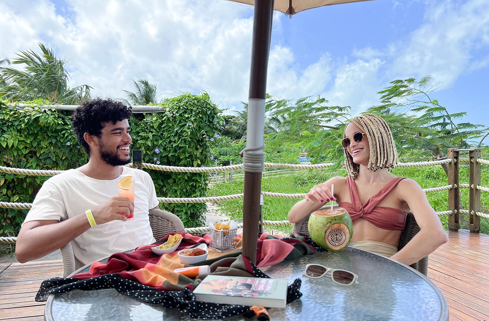  Casal sorrindo e relaxando em uma mesa redonda ao ar livre, com drinks e petiscos. O homem está com um coquetel e a mulher com um coco natural, com vegetação e céu azul ao fundo.