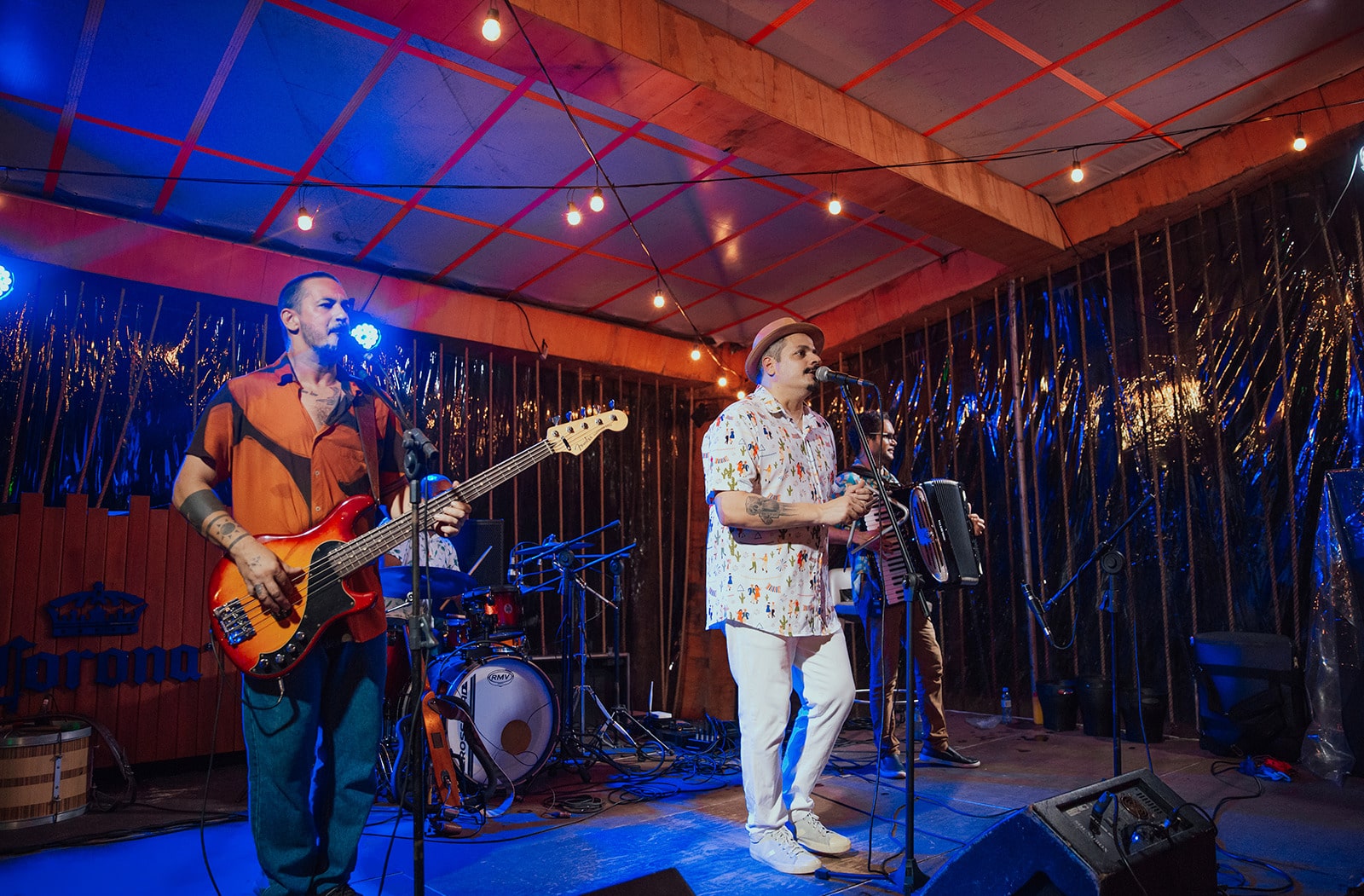 Uma banda de quatro músicos se apresentando em um palco iluminado. Os integrantes tocam baixo, acordeão e bateria. O cenário tem luzes azuis e amarelas.