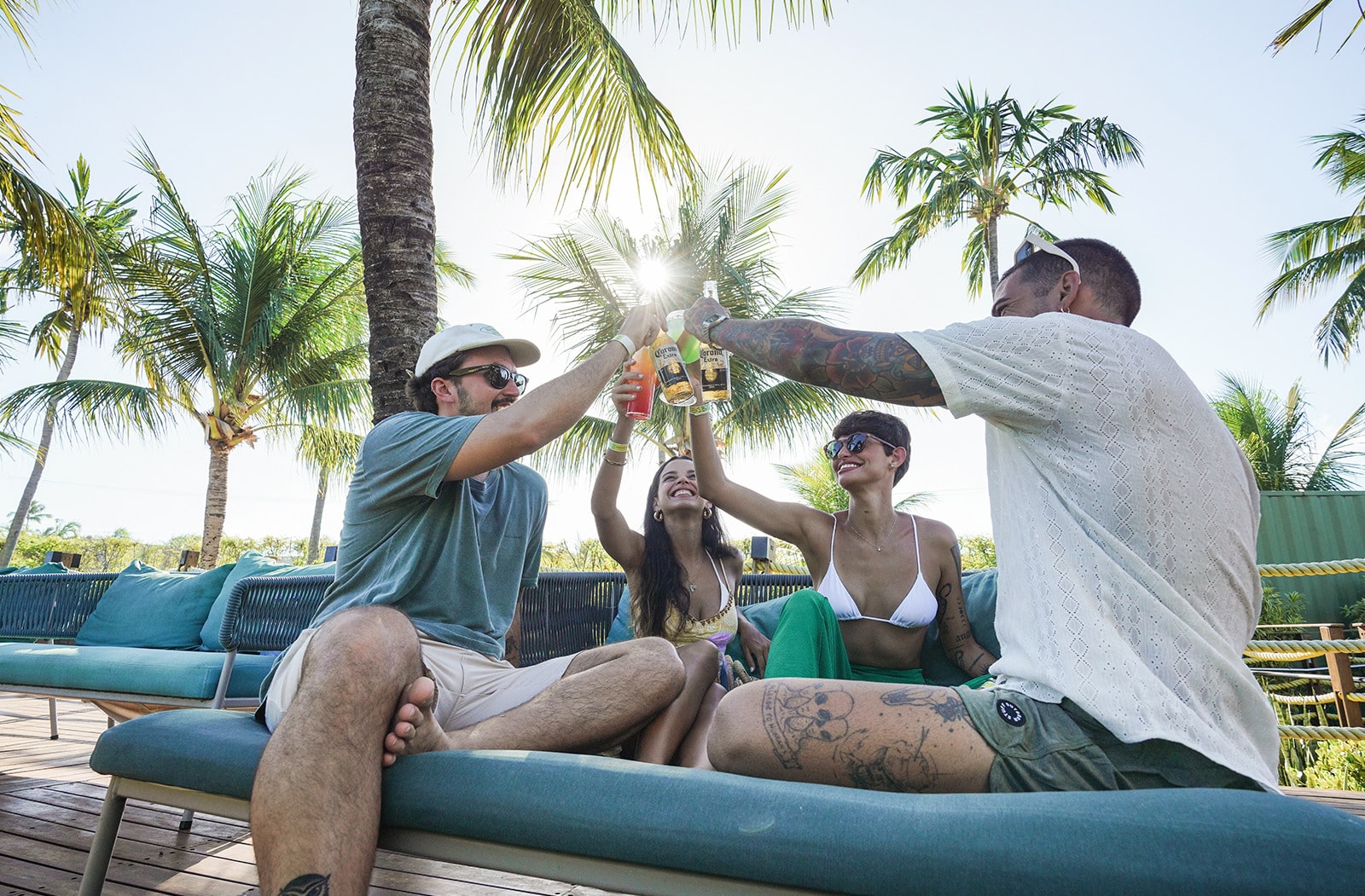 Quatro amigos brindando com drinks e cerveja em um lounge externo, sentados em grandes cadeiras de praia. A imagem mostra o sol brilhando por trás das palmeiras.