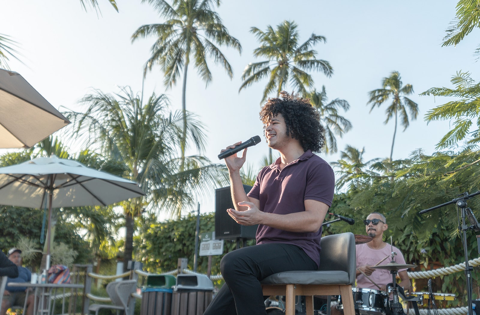 Cantor cantando, sentado em uma cadeira com microfone na mão, durante um show de música ao vivo ao ar livre. Outro músico está atrás dele, tocando bateria, com palmeiras ao fundo e um céu sem nuvens.