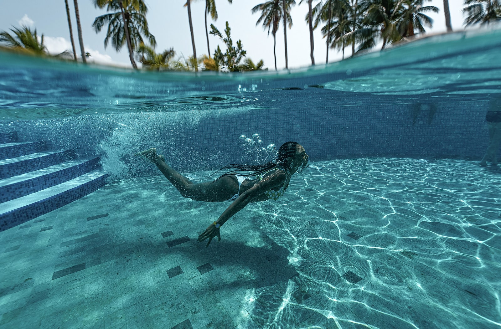  Mulher mergulhando em uma piscina em uma foto subaquática e de superfície. A imagem mostra a água cristalina com bolhas, com coqueiros e um céu azul ao fundo.