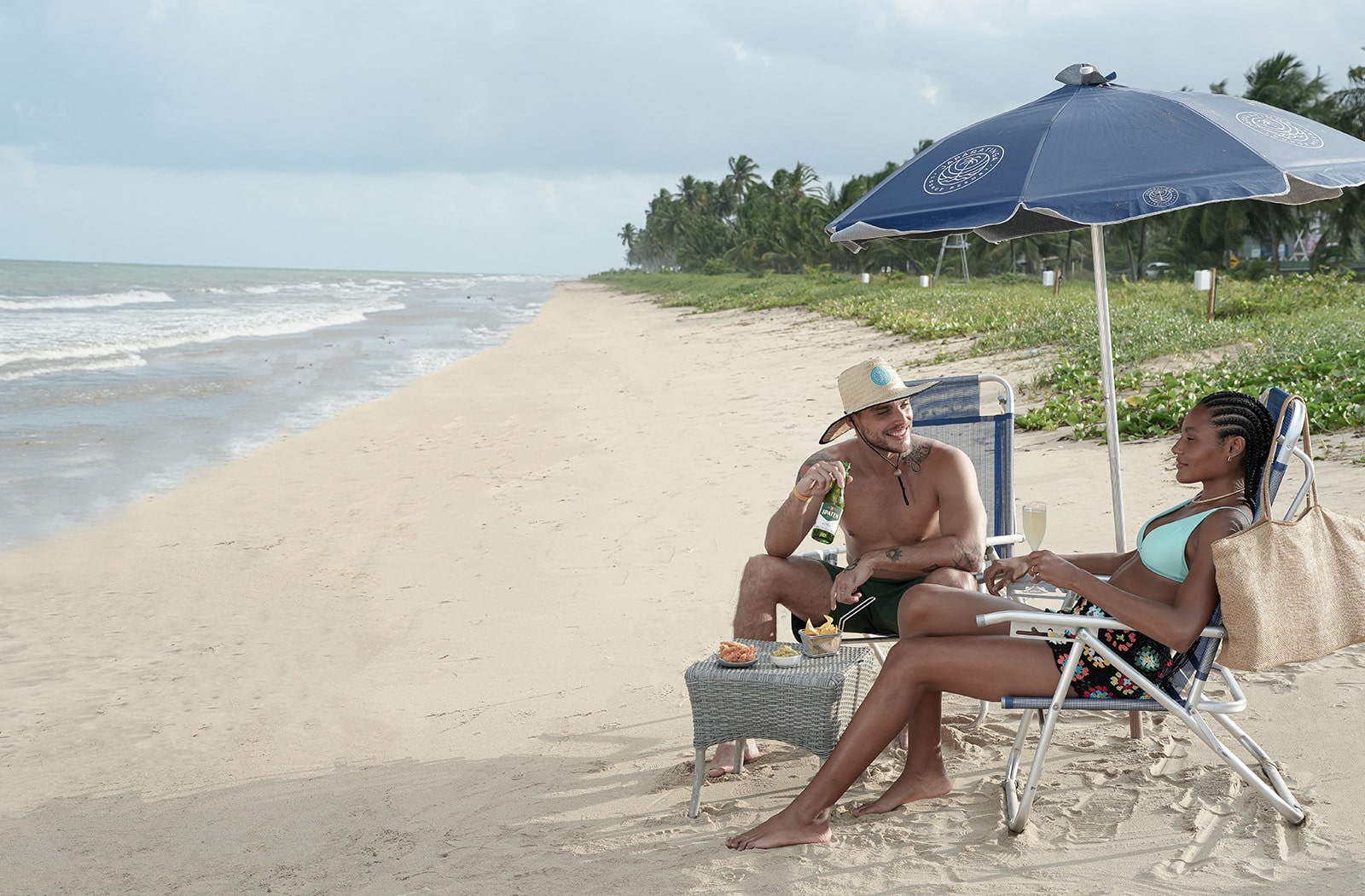 Casal relaxando em cadeiras de praia sob um guarda-sol azul. Eles estão na beira do mar com drinks e petiscos, com palmeiras e céu azul ao fundo.