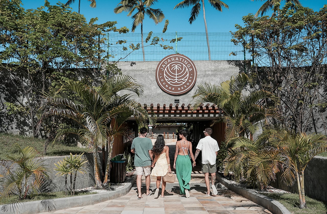  Casal de amigos de costas caminhando em direção à entrada do túnel de Japaratinga Lounge Resort, com céu azul e muito verde