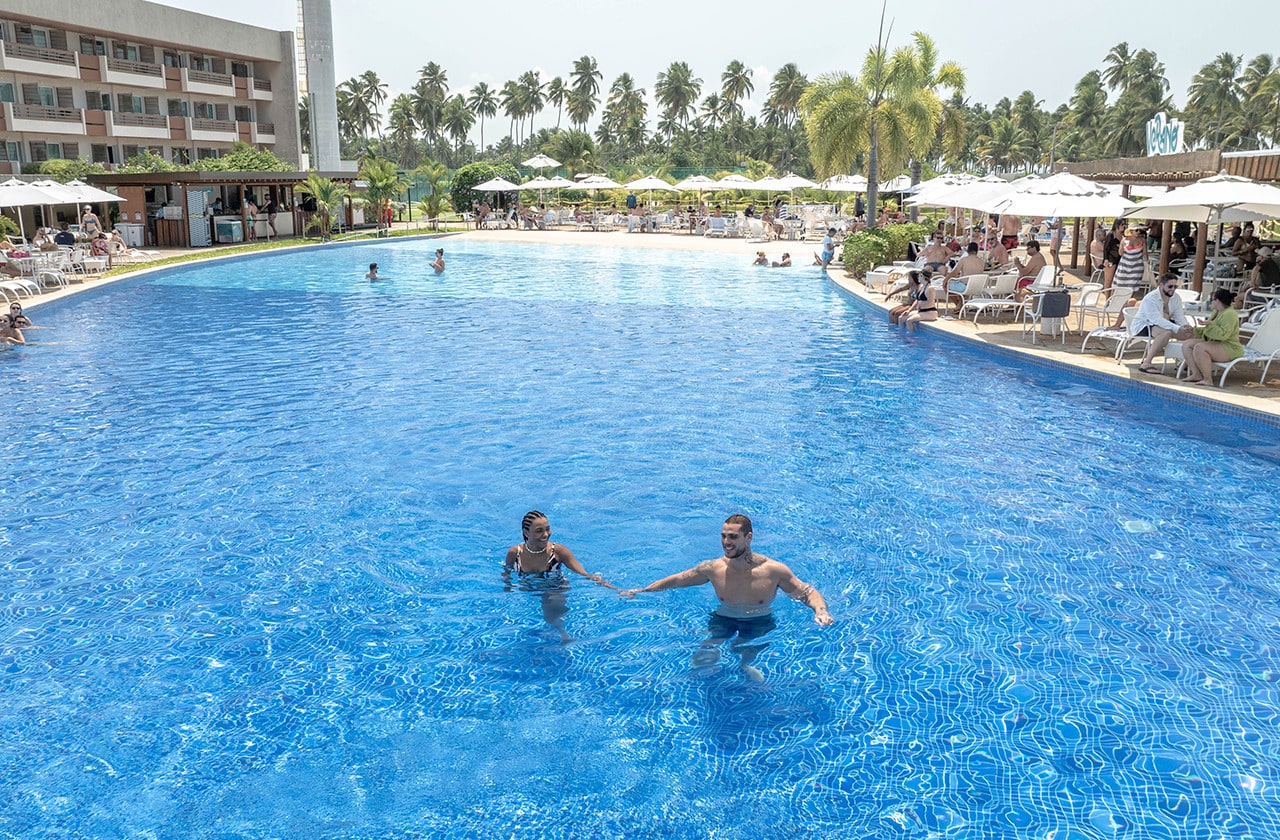 Casal de mãos dadas curtindo a piscina principal do Japaratinga Lounge Resort, com outros hóspedes relaxando nas espreguiçadeiras ao redor e coqueiros ao fundo.